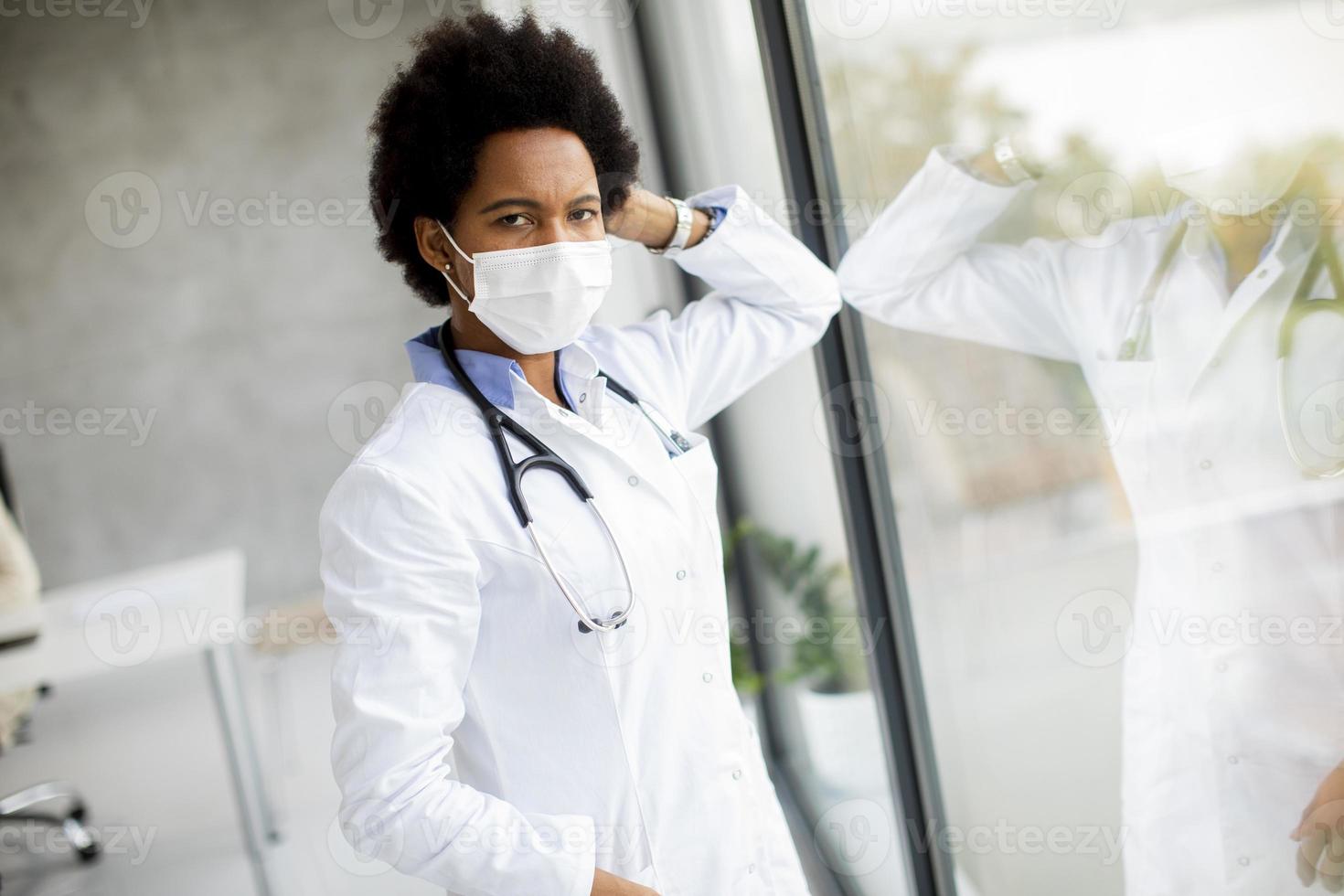 Doktor mit Maske, die sich gegen Fenster lehnt und Kamera betrachtet foto