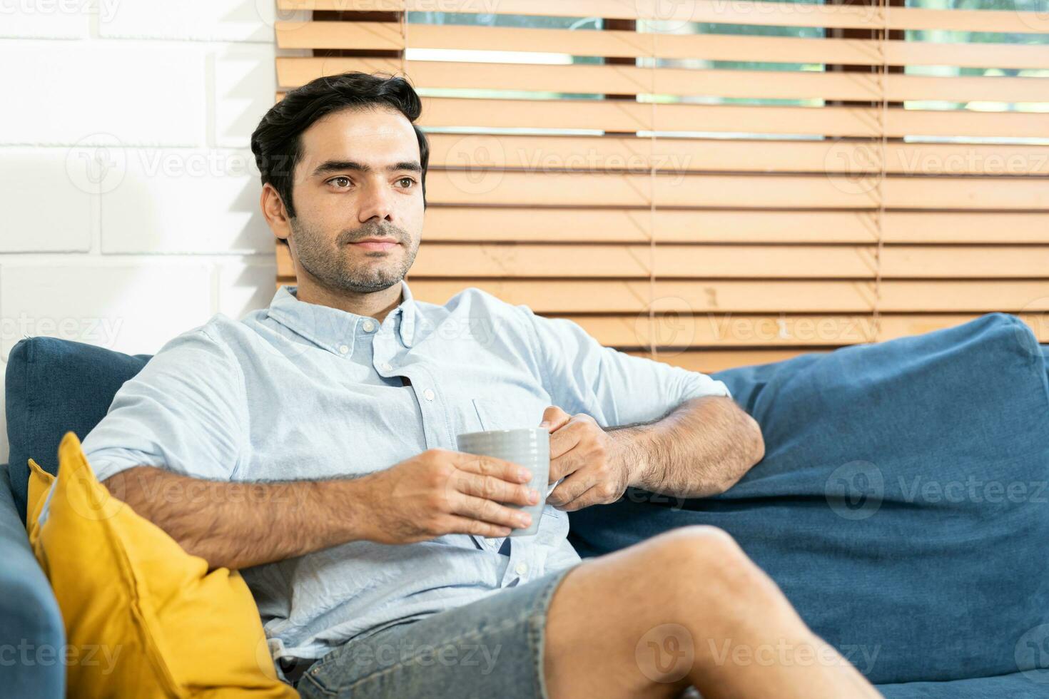 sich ausruhen Konzept. glücklich Kerl Trinken Kaffee Sitzung auf komfortabel Couch beim Zuhause im Leben Zimmer. ein lächelnd jung bärtig Mann Trinken Tee von ein Becher beim das Leben Zimmer. foto