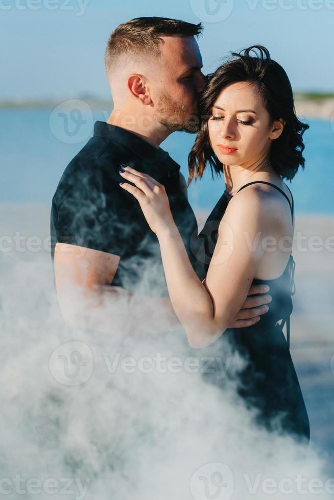 Mann und ein Mädchen in schwarzer Kleidung umarmen sich in weißem Rauch foto