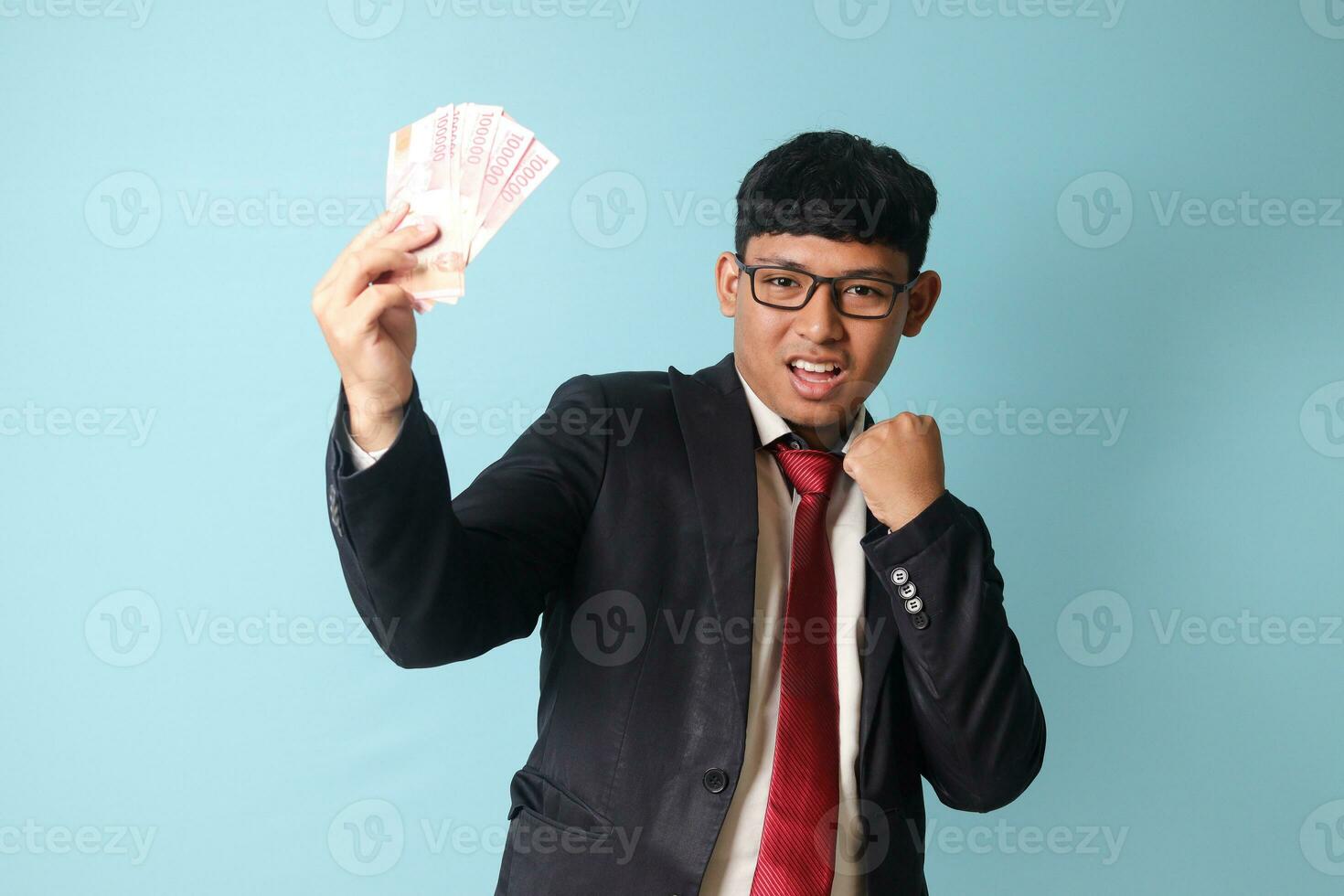 Porträt von jung asiatisch Geschäft Mann im beiläufig passen mit glücklich Ausdrücke halten tausend Rupien und feiern. isoliert Bild auf Blau Hintergrund foto