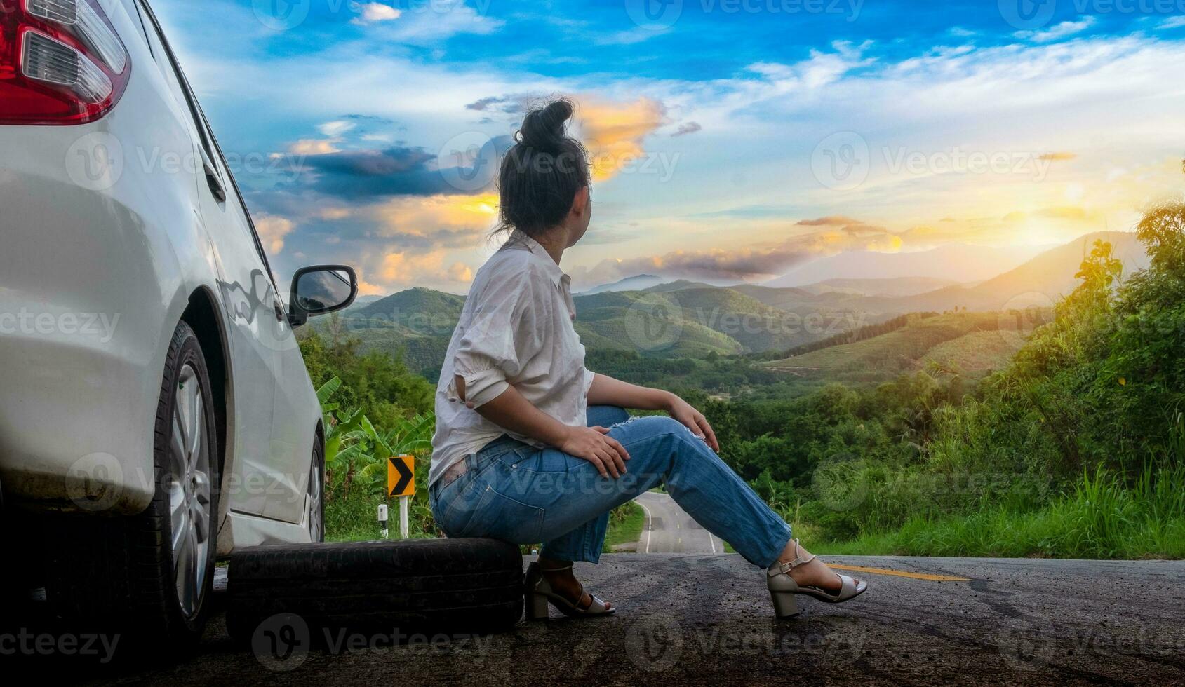 junge schöne Dame, die nahe einem Auto sitzt, um auf der öffentlichen Straße in einem Waldgebiet mit einem Berg- und Himmelhintergrund um Hilfe zu rufen foto