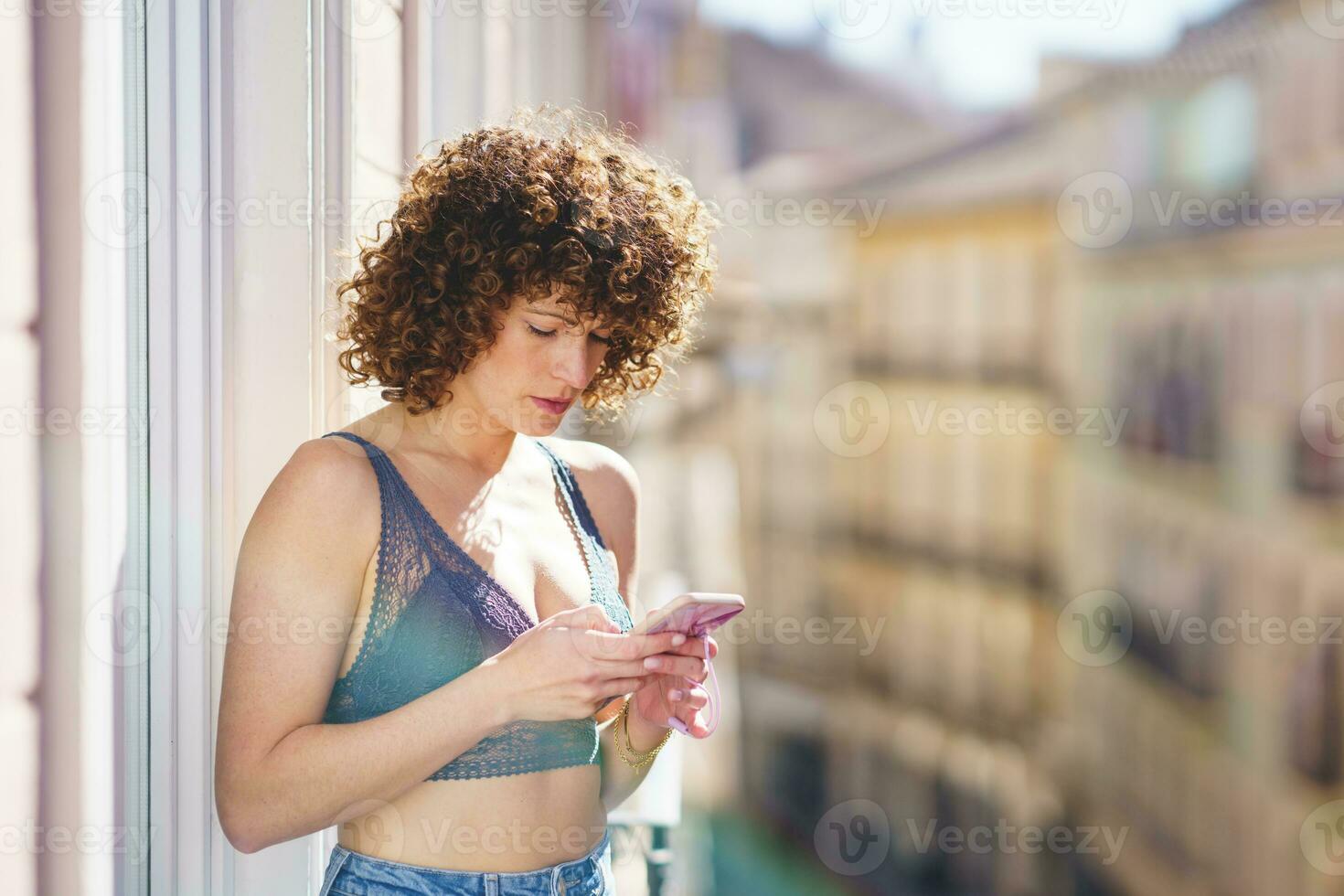 jung weiblich im BH und Jeans mit lockig Haar während Surfen Smartphone foto