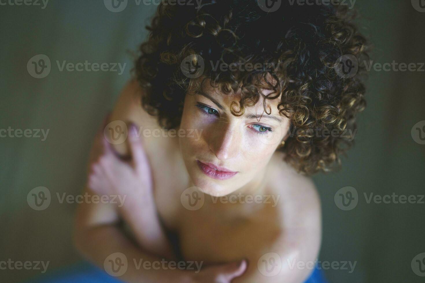 hoch Winkel von Frau mit lockig Haar, mit ihr nackt Schultern Blick durch das Fenster foto