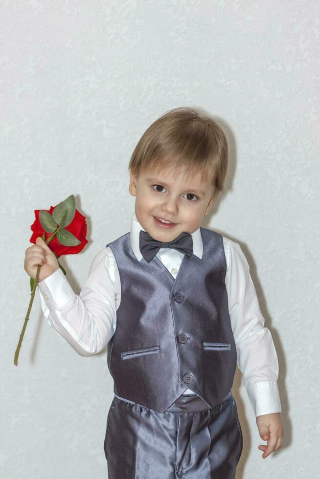 ein wenig Junge hält und Hände Über ein rot Rose, das Konzept von das Valentinstag Tag Thema. Porträt von ein süß Junge im ein passen mit ein Bogen binden. foto