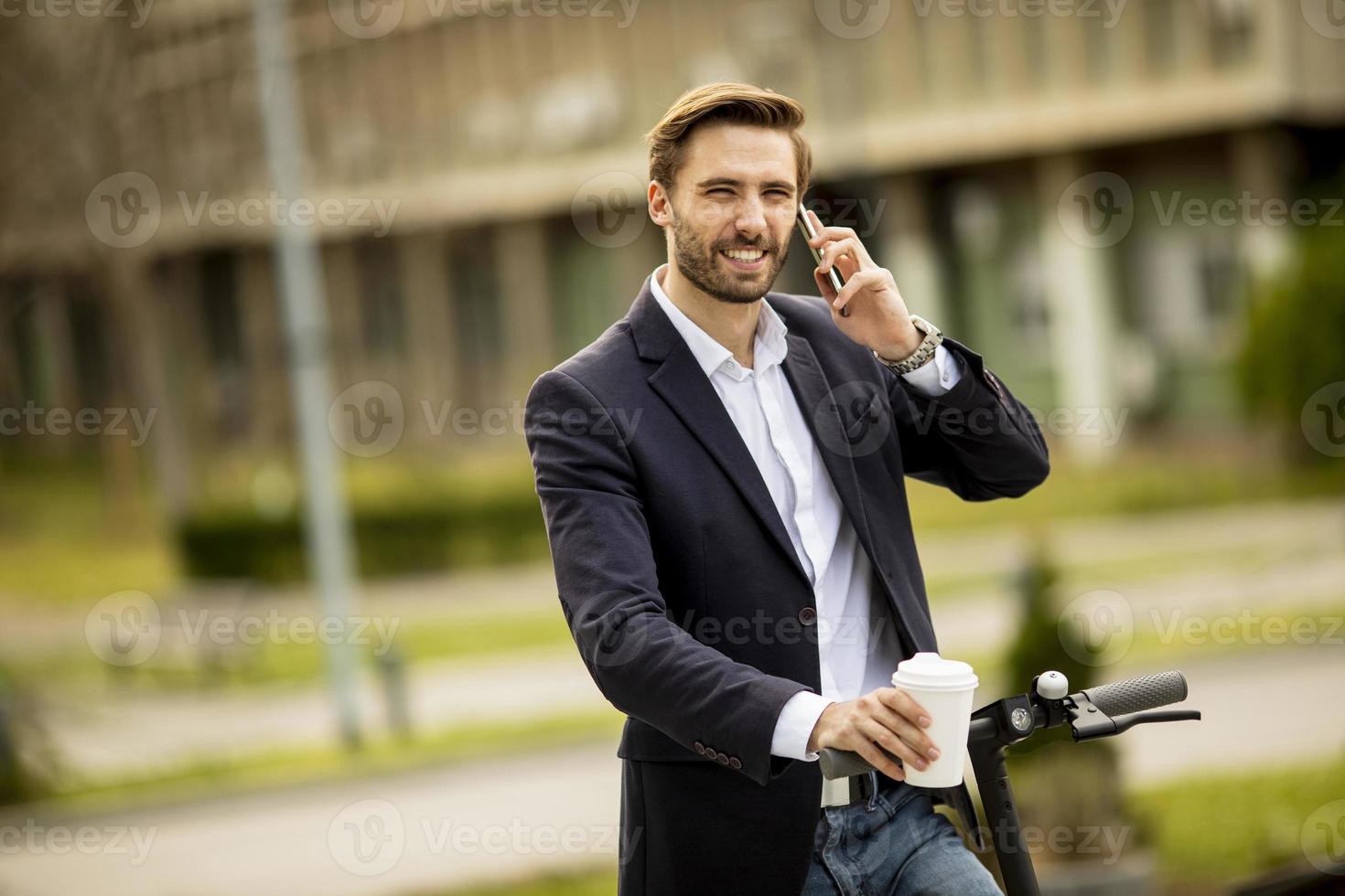 Gelegenheitsgeschäftsmann, der einen Kaffee hält und auf einem Roller am Telefon spricht foto