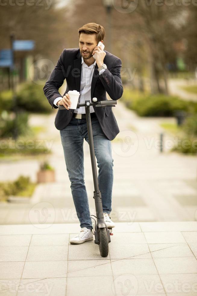 Gelegenheitsgeschäftsmann, der einen Kaffee hält und auf einem Telefon spricht, während er auf einem Roller steht foto