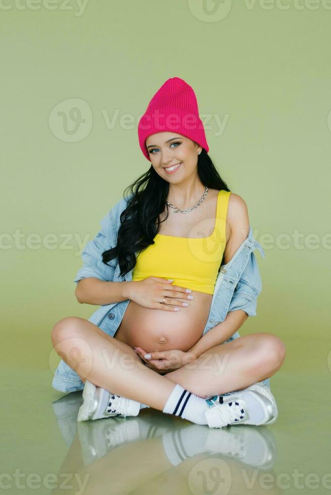 ein jung lächelnd schwanger Frau im ein Rosa Hut und hell stilvoll Kleider hält ihr Hände auf ihr Bauch auf ein Grün Olive Hintergrund foto