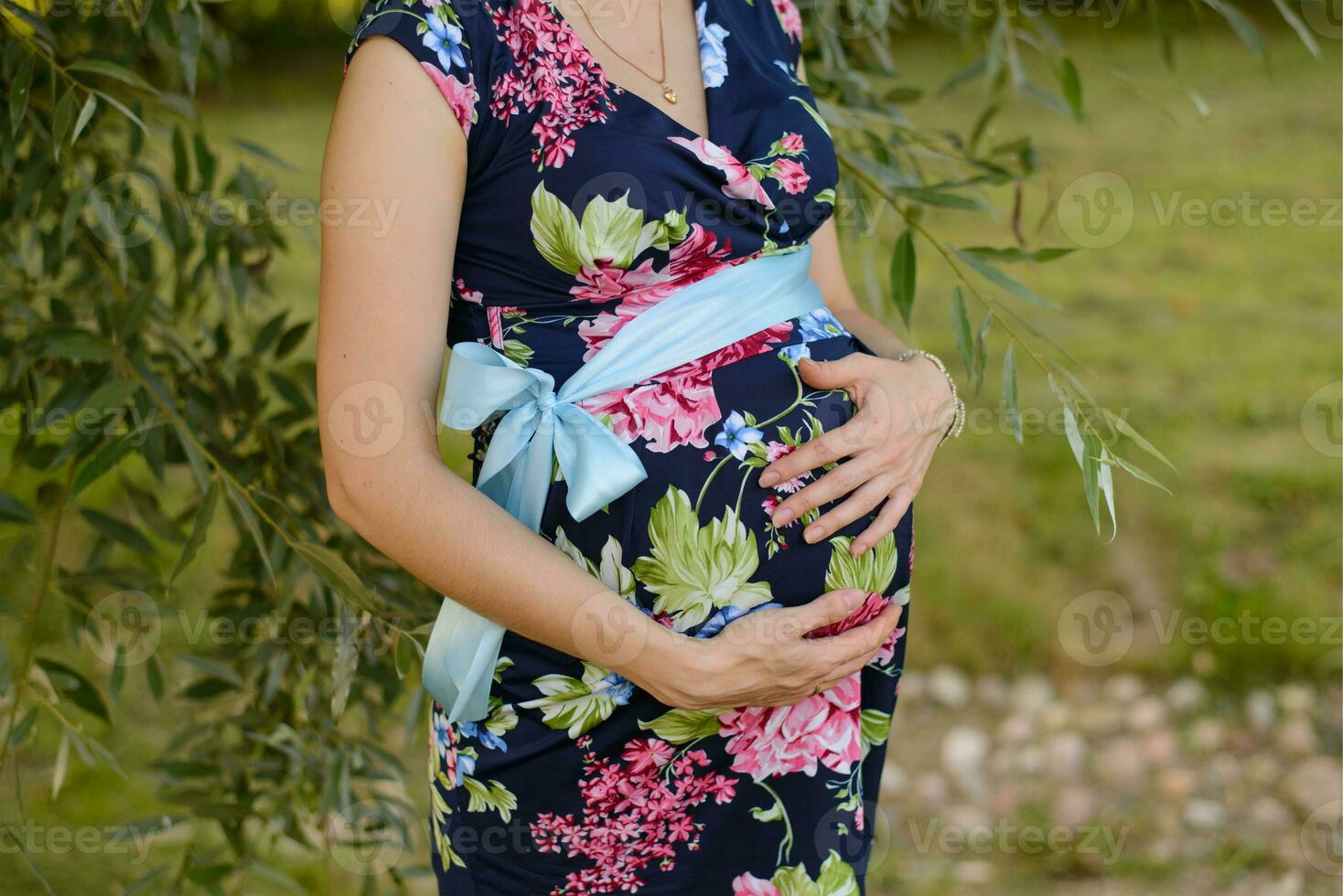 ein schwanger Frau im ein hell farbig Kleid hält ihr Hände auf ihr Bauch während warten zum ein Baby foto