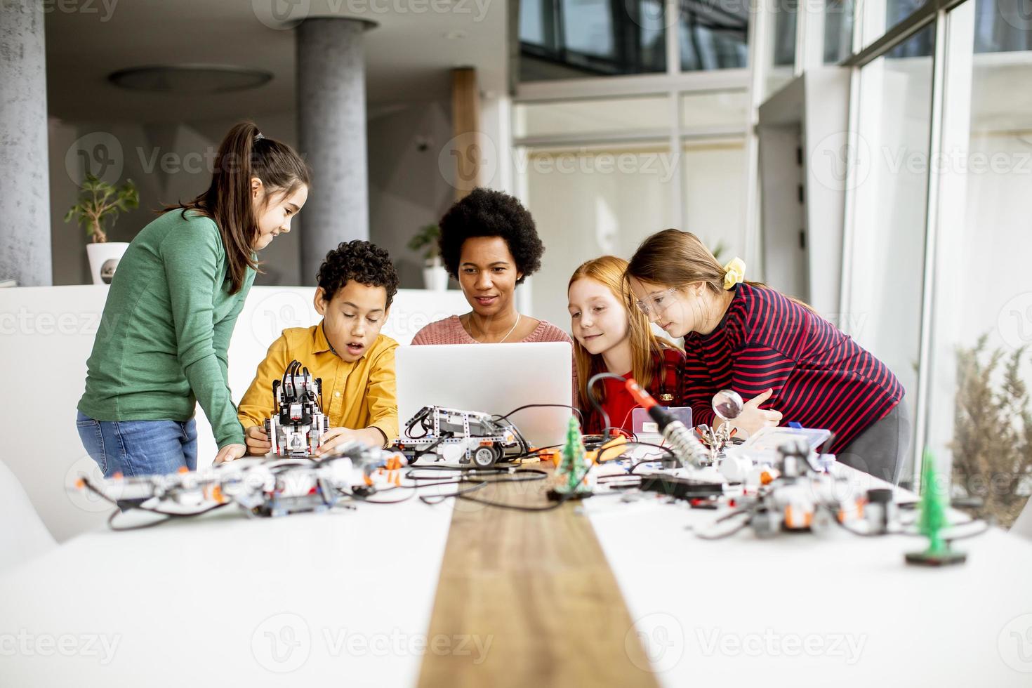 glückliche Kinder mit ihrer afroamerikanischen Wissenschaftslehrerin mit Laptop-Programmierung von elektrischem Spielzeug und Robotern im Robotik-Klassenzimmer foto