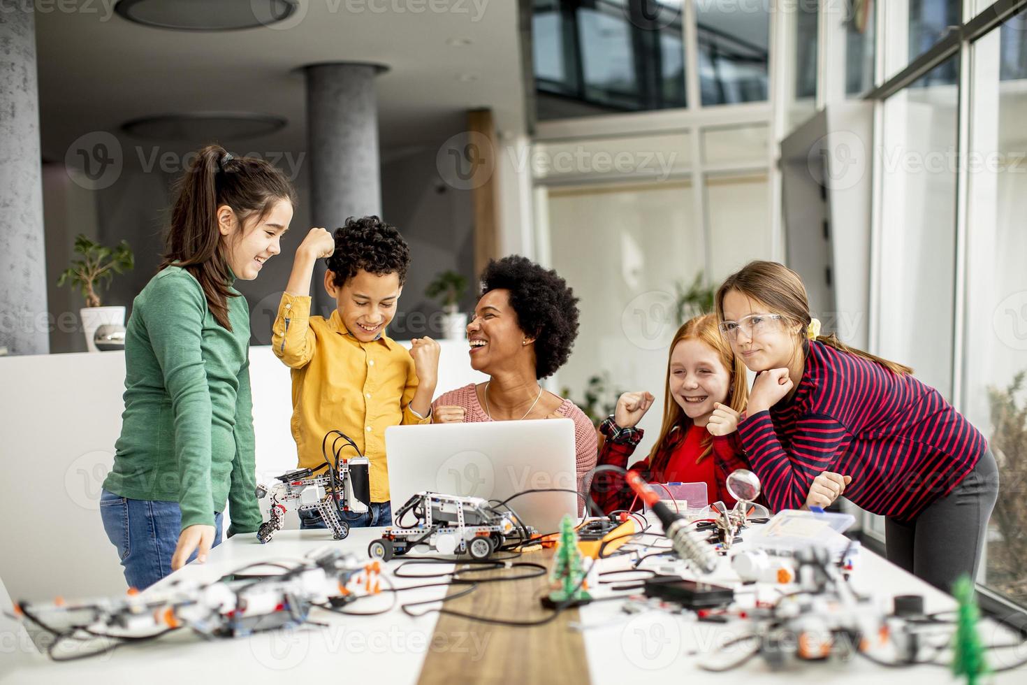 glückliche Kinder mit ihrer afroamerikanischen Wissenschaftslehrerin mit Laptop-Programmierung von elektrischem Spielzeug und Robotern im Robotik-Klassenzimmer foto