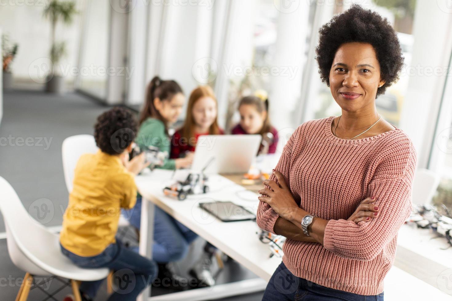 Afroamerikaner-Wissenschaftslehrerin mit Gruppe von Kindern, die elektrisches Spielzeug und Roboter am Robotik-Klassenzimmer programmieren foto