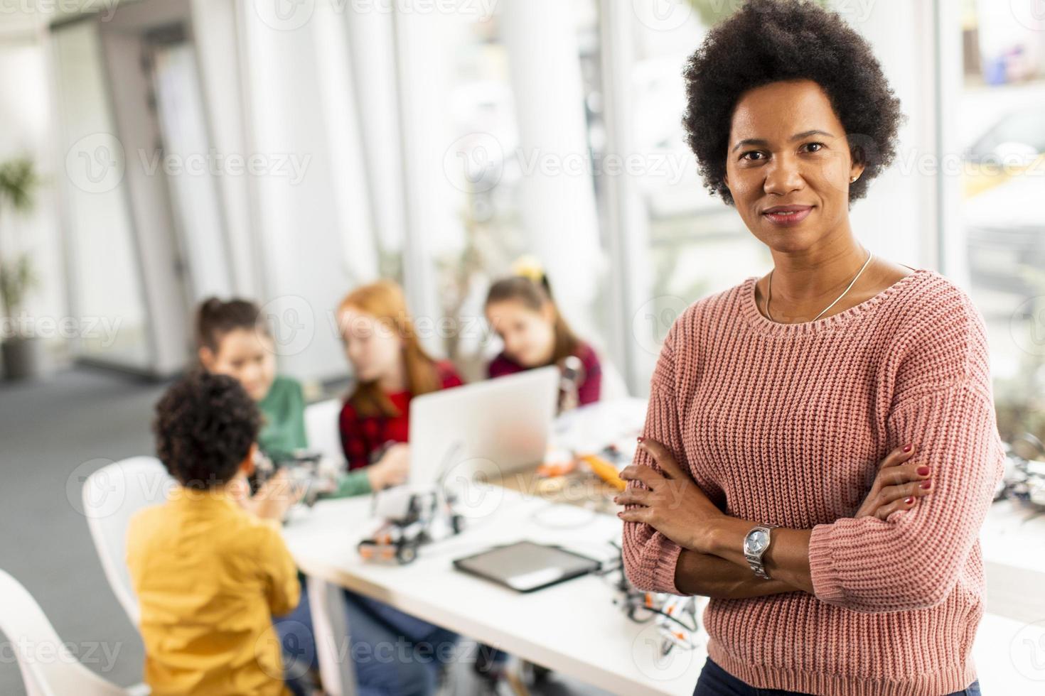 Afroamerikaner-Wissenschaftslehrerin mit Gruppe von Kindern, die elektrisches Spielzeug und Roboter am Robotik-Klassenzimmer programmieren foto