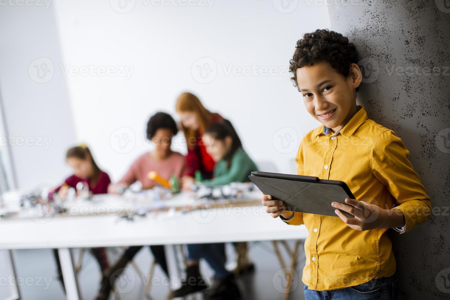 süßer kleiner Junge, der vor Kindern steht, die elektrisches Spielzeug und Roboter am Robotikklassenzimmer programmieren foto