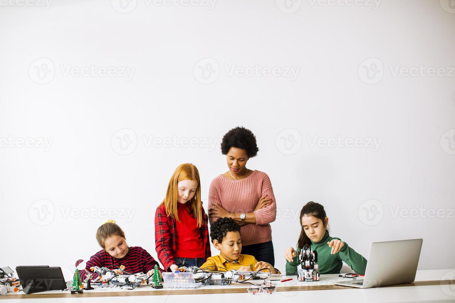 glückliche Kinder mit ihrer afroamerikanischen Wissenschaftslehrerin, die elektrisches Spielzeug und Roboter im Robotikklassenzimmer programmiert foto