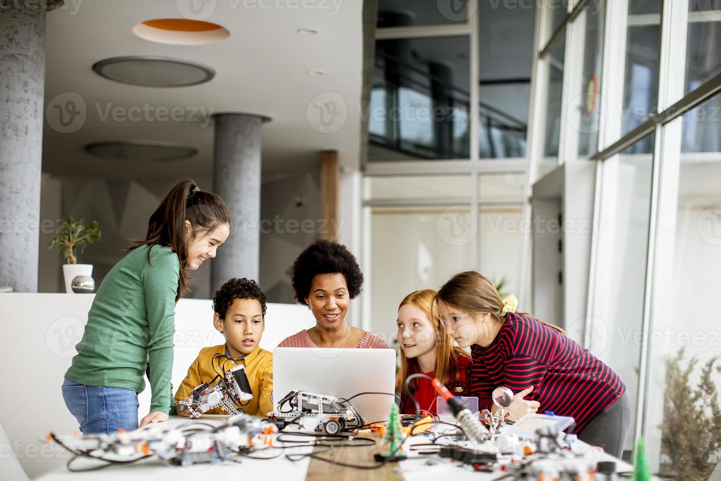 glückliche Kinder mit ihrer afroamerikanischen Wissenschaftslehrerin mit Laptop-Programmierung von elektrischem Spielzeug und Robotern im Robotik-Klassenzimmer foto