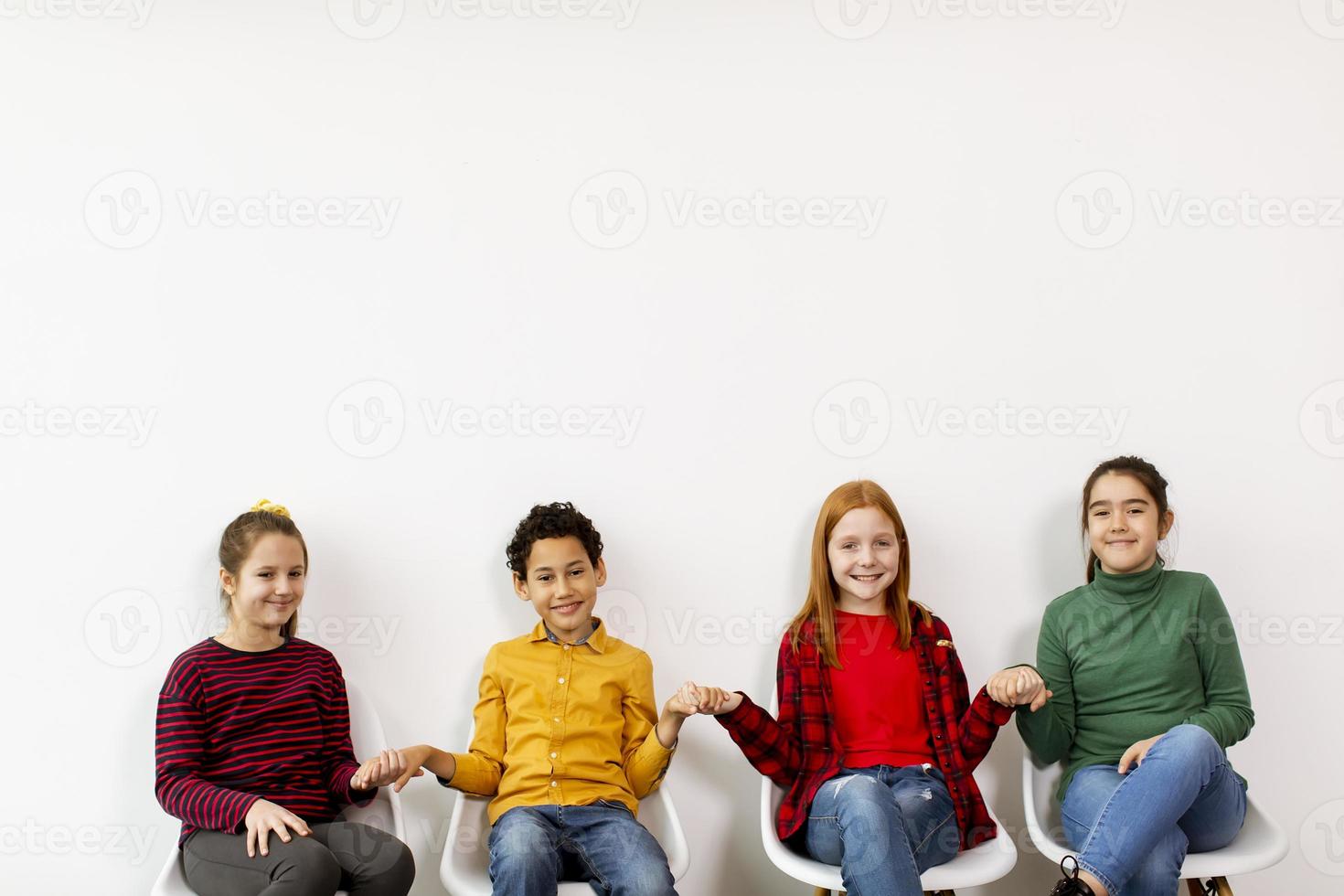 Porträt der niedlichen kleinen Kinder in den Jeans, die in Stühlen gegen weiße Wand sitzen foto