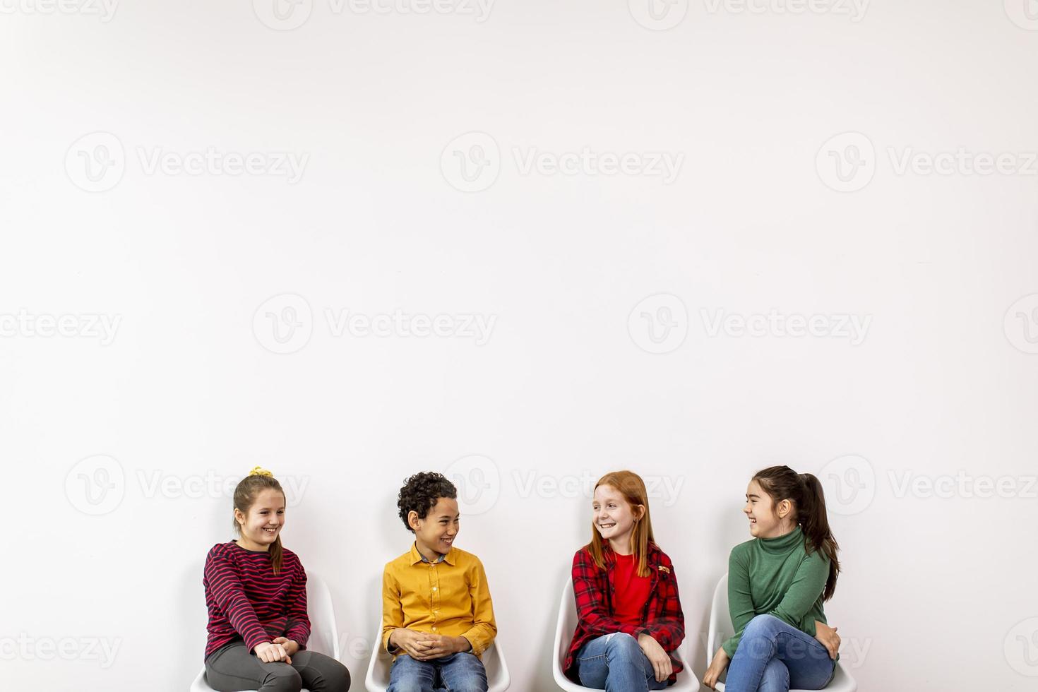 Porträt der niedlichen kleinen Kinder in den Jeans, die in Stühlen gegen weiße Wand sitzen foto