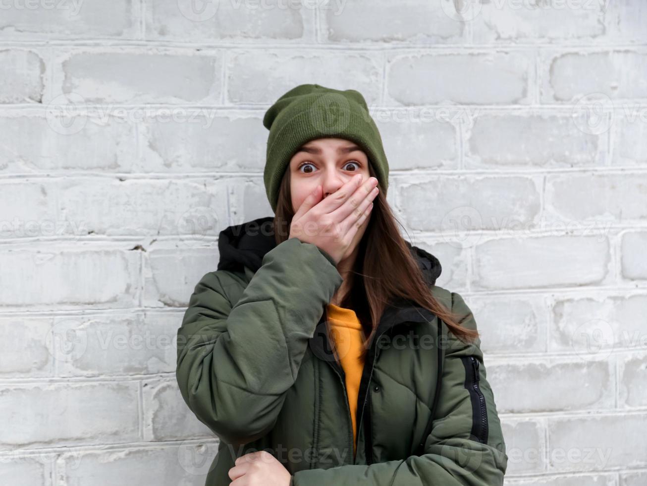 Porträt eines schönen schockierten Mädchens, das ihren Mund mit der Hand in einem gelben Pullover und einem Khakihut schließt, die nahe einer weißen Backsteinmauer stehen foto