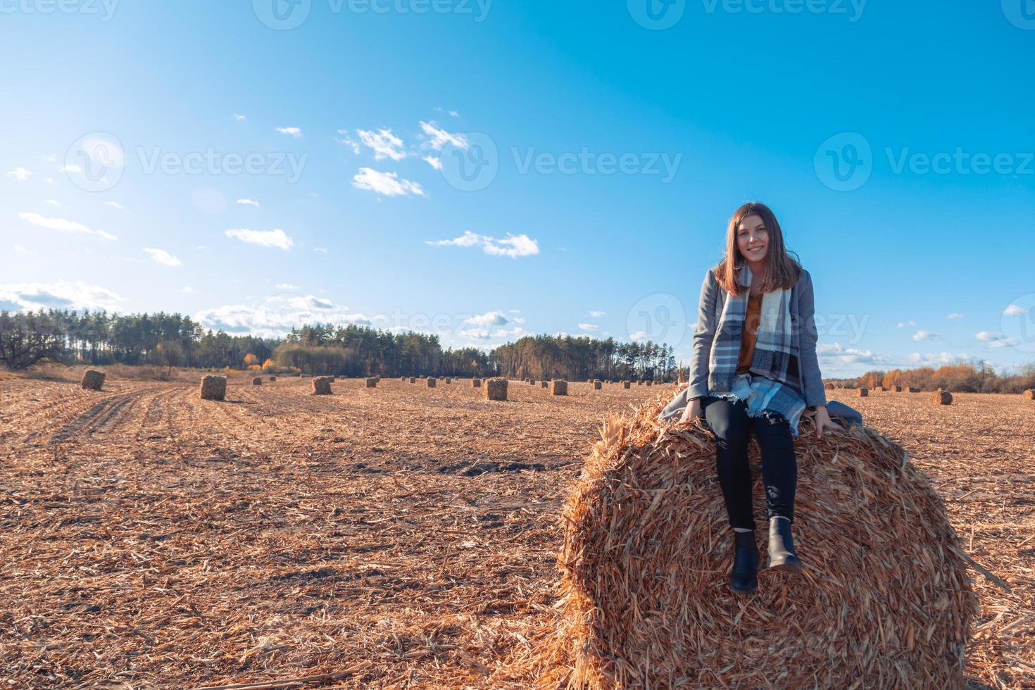 Ein Mädchen von europäischem Aussehen in einem grauen Mantel steht auf einem Feld in der Nähe eines größeren Ballens mit Heu vor einem blauen Himmel foto