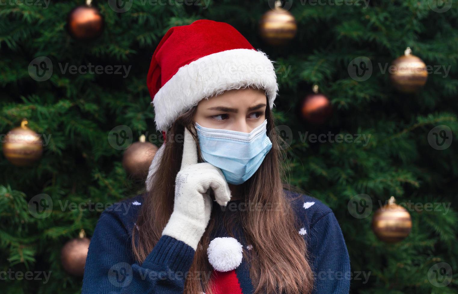 Nahaufnahmeporträt der Frau, die einen Weihnachtsmannhut und eine medizinische Maske mit Emotion trägt foto