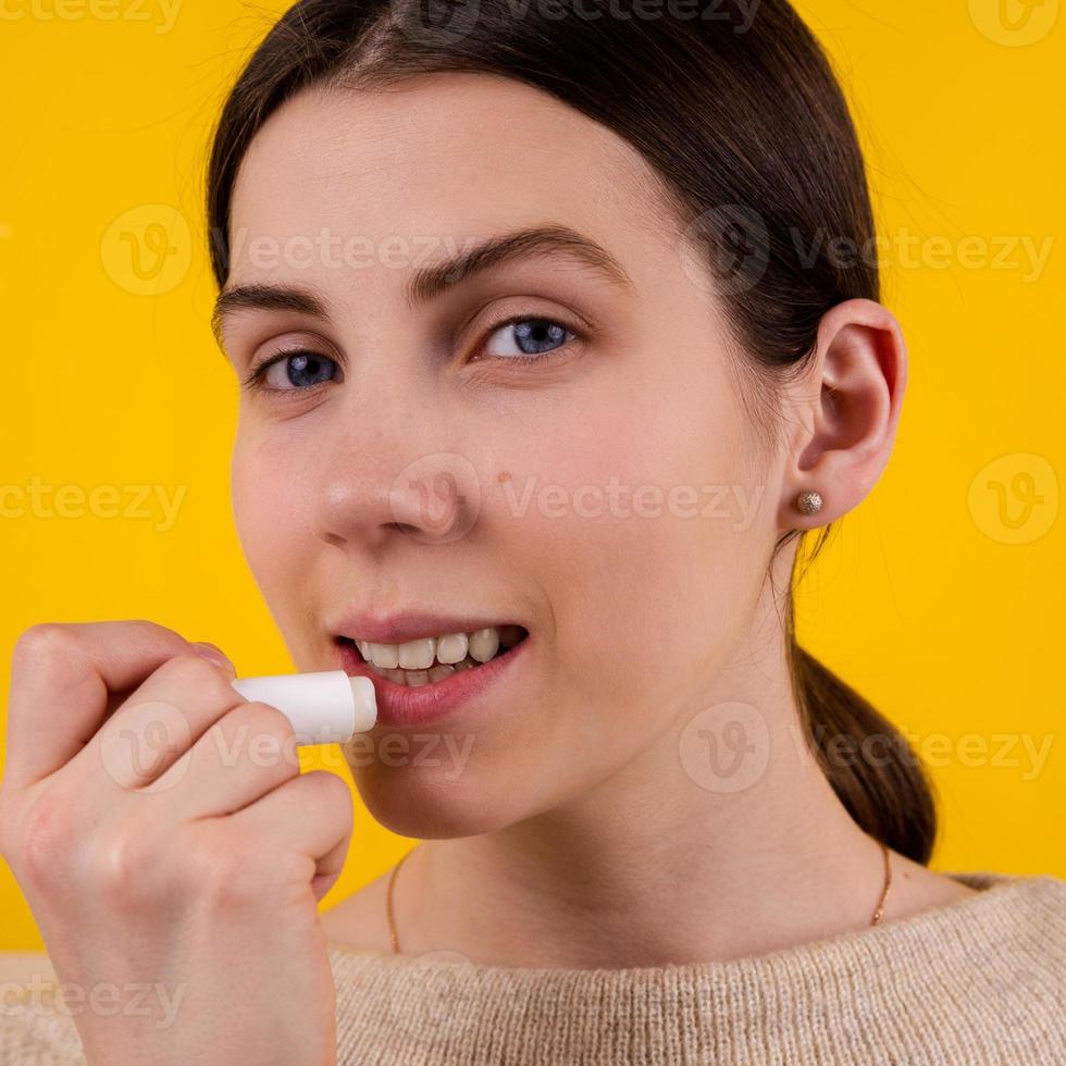 attraktive lächelnde junge Frau, die hygienischen Lippenstift auf gelbem Hintergrund verwendet foto