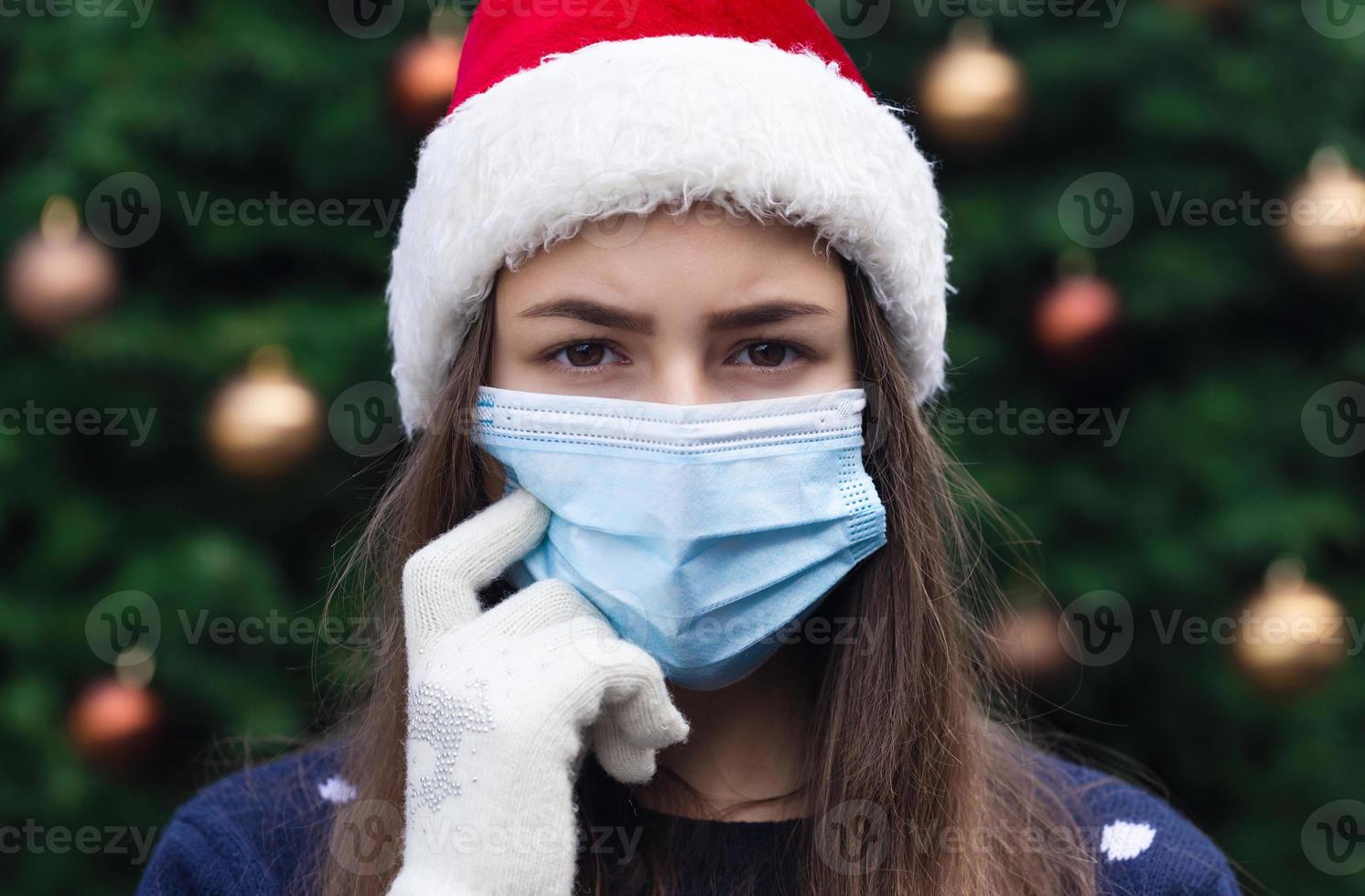 Nahaufnahmeporträt der Frau, die einen Weihnachtsmannhut und eine medizinische Maske mit Emotion trägt foto