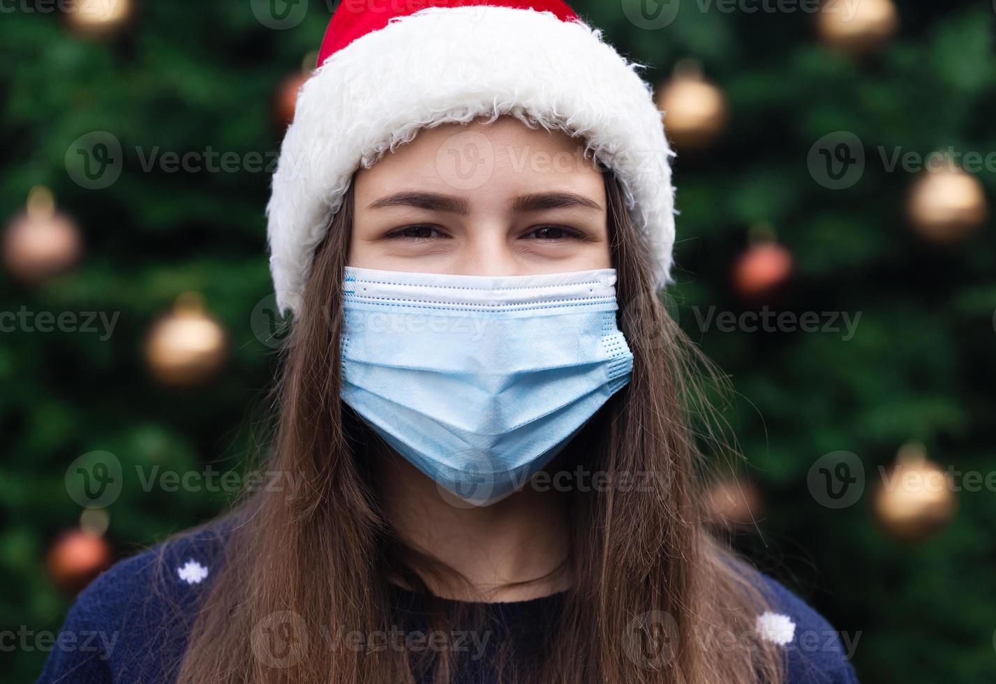 Nahaufnahmeporträt der Frau, die einen Weihnachtsmannhut und eine medizinische Maske mit Emotion trägt foto