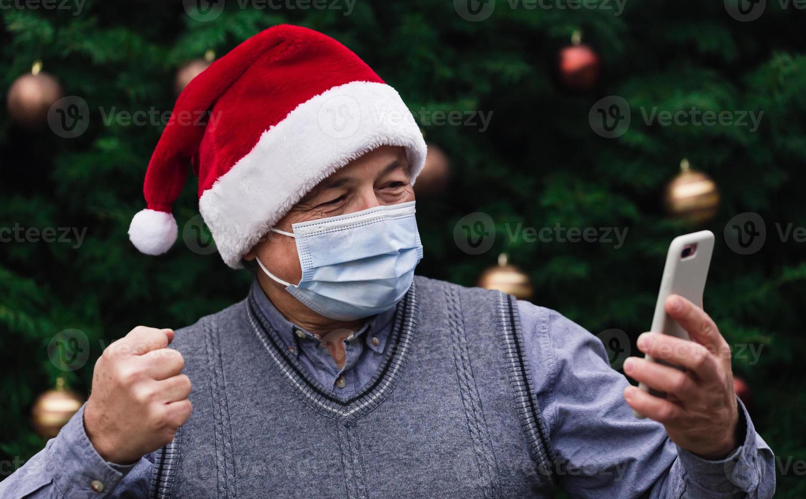 Nahaufnahmeporträt des älteren Mannes, der einen Weihnachtsmannhut und eine medizinische Maske mit Emotion trägt foto