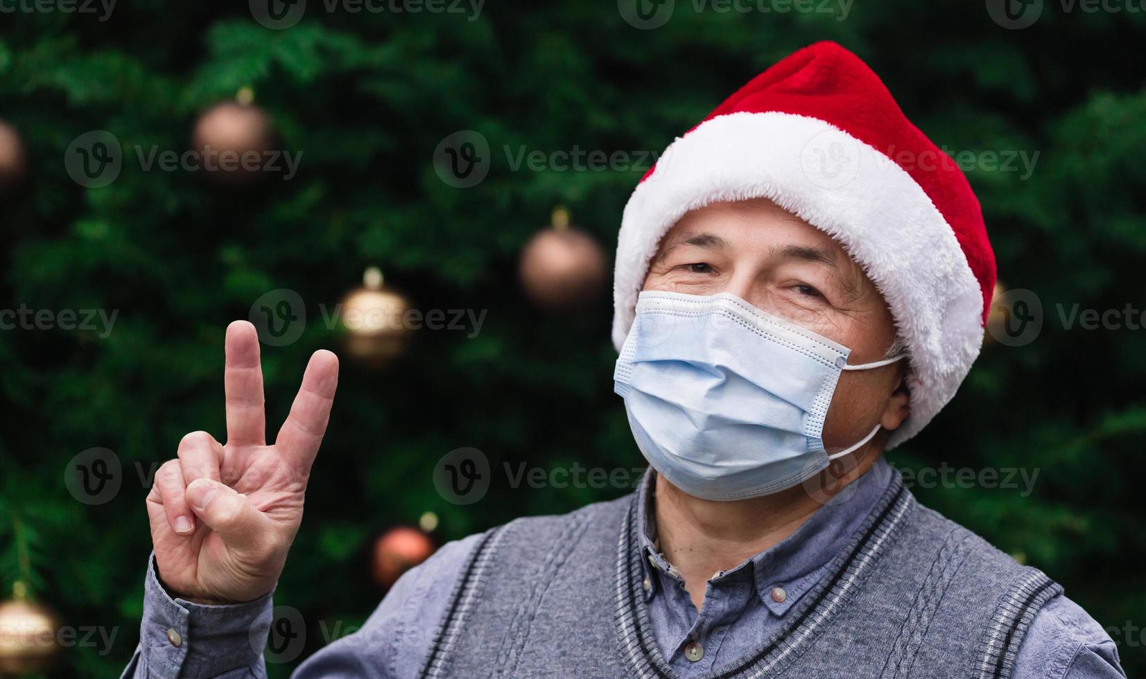 Nahaufnahmeporträt des älteren Mannes, der einen Weihnachtsmannhut und eine medizinische Maske mit Emotion trägt foto