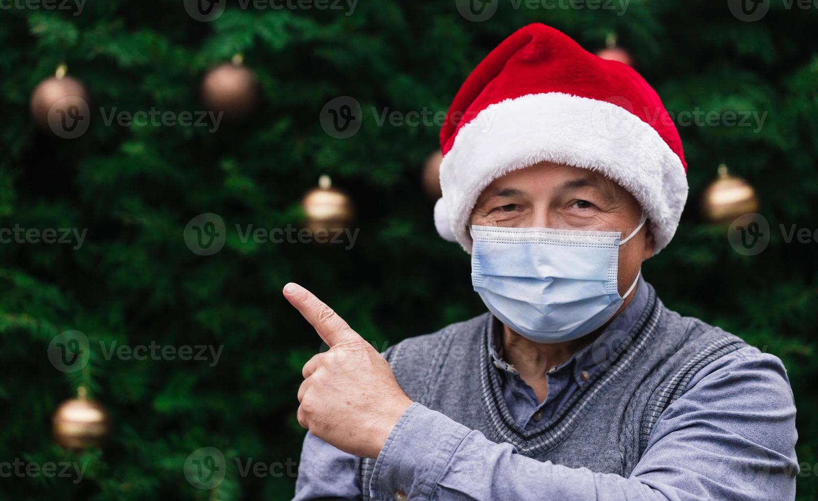 Nahaufnahmeporträt des älteren Mannes, der einen Weihnachtsmannhut und eine medizinische Maske mit Emotion trägt foto