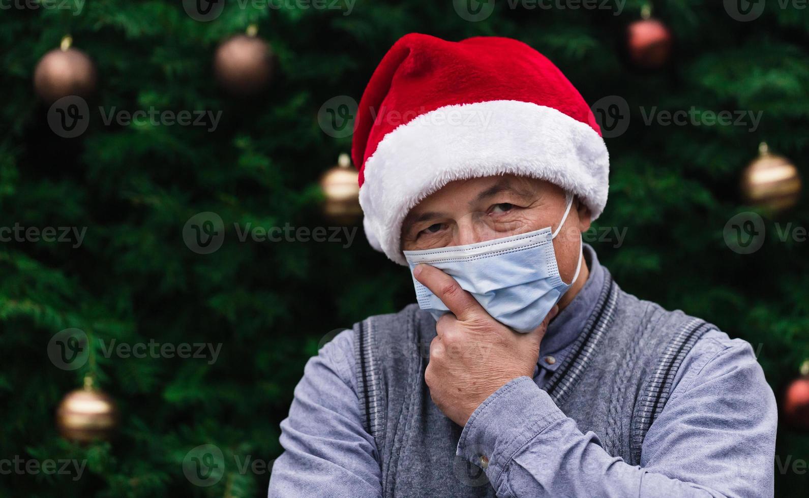 Nahaufnahmeporträt des älteren Mannes, der einen Weihnachtsmannhut und eine medizinische Maske mit Emotion trägt foto