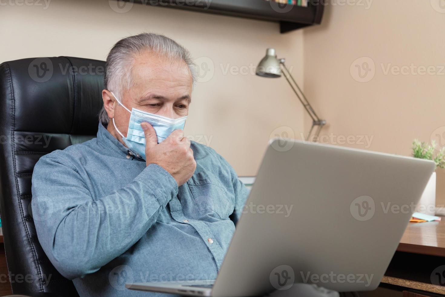 älterer Mann in der Gesichtsmaske, die zu Hause am Laptop arbeitet oder kommuniziert foto