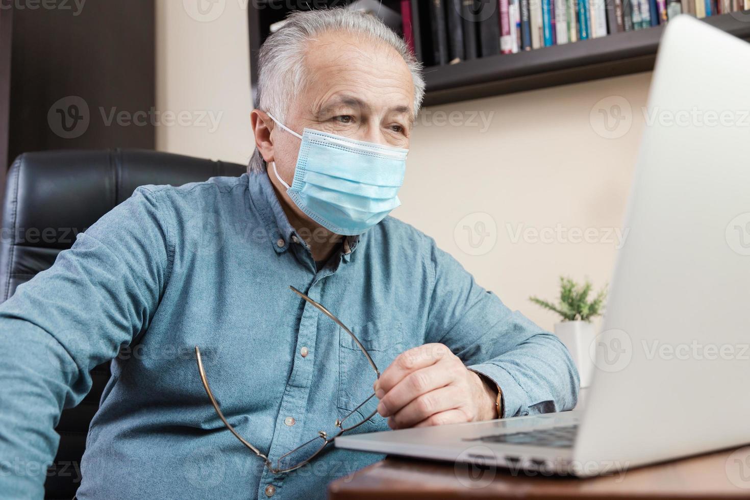 älterer Mann in der Gesichtsmaske, die zu Hause am Laptop arbeitet oder kommuniziert foto