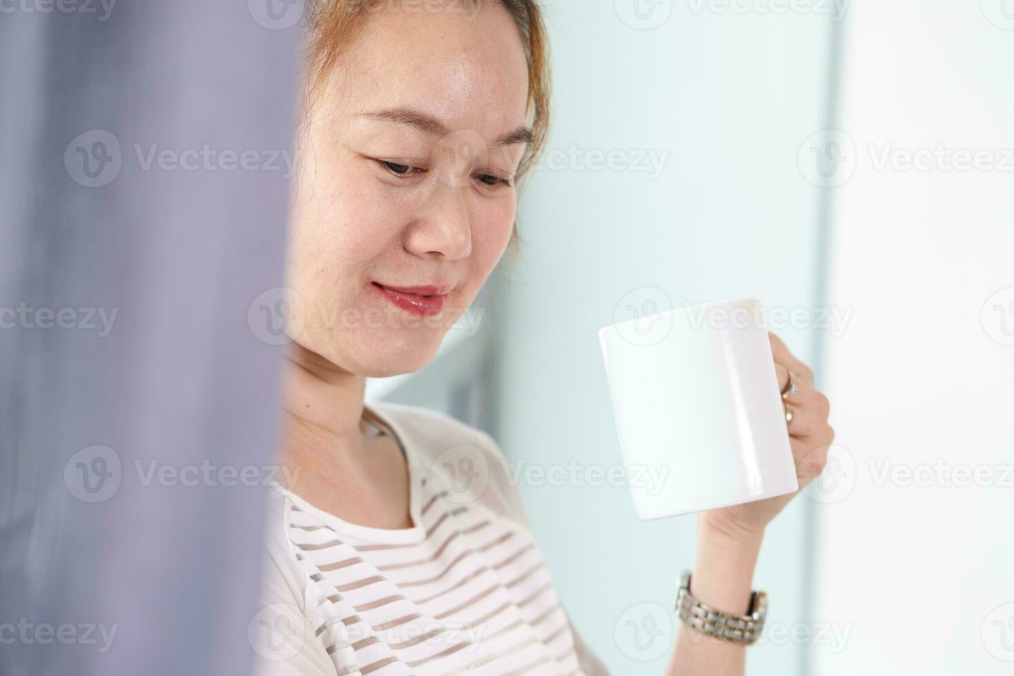 schön attraktiv Mitte Alter Süd-Ost asiatisch Frau Stehen Nächster zu Tageslicht Fenster Vorhang trinken Kaffee von Tasse suchen draußen foto