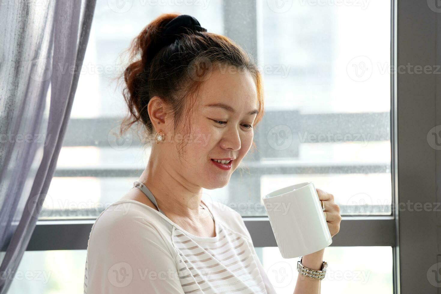 schön attraktiv Mitte Alter Süd-Ost asiatisch Frau Stehen Nächster zu Tageslicht Fenster Vorhang trinken Kaffee von Tasse suchen Nieder foto