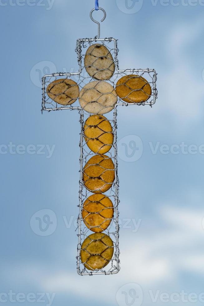 religiöses Kreuz aus Kieselsteinen und Drahtgeflecht, das vor dem blauen Himmel hängt foto