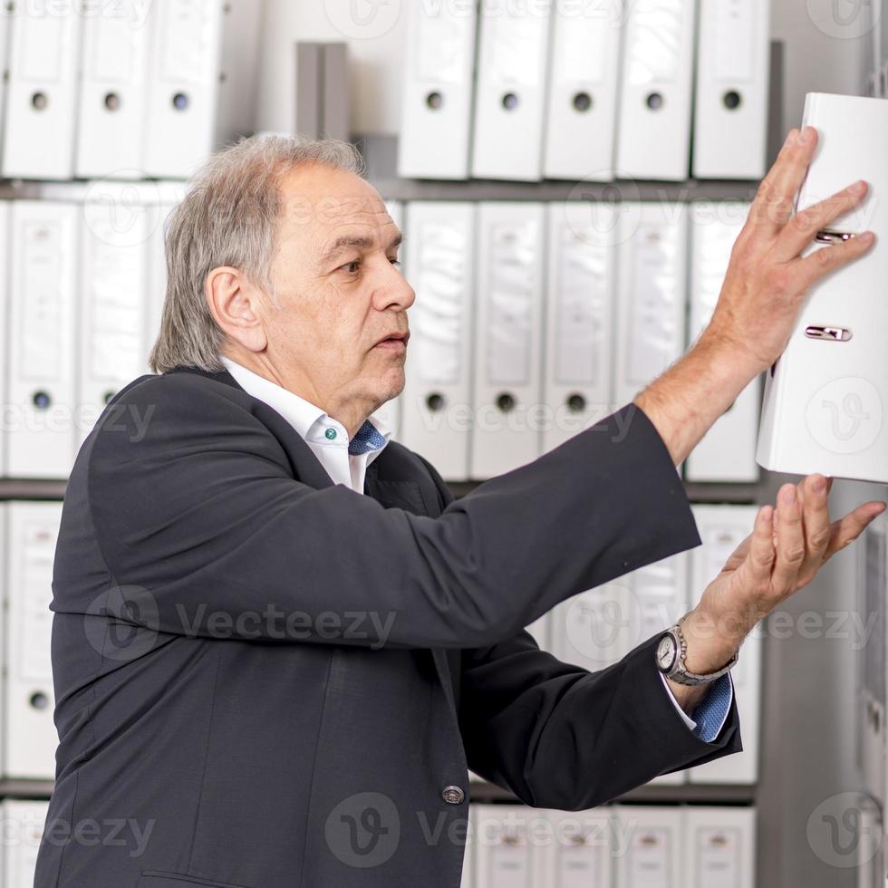 Mann mittleren Alters mit weißem Hemd steht vor einer Regalwand mit Dokumenten foto
