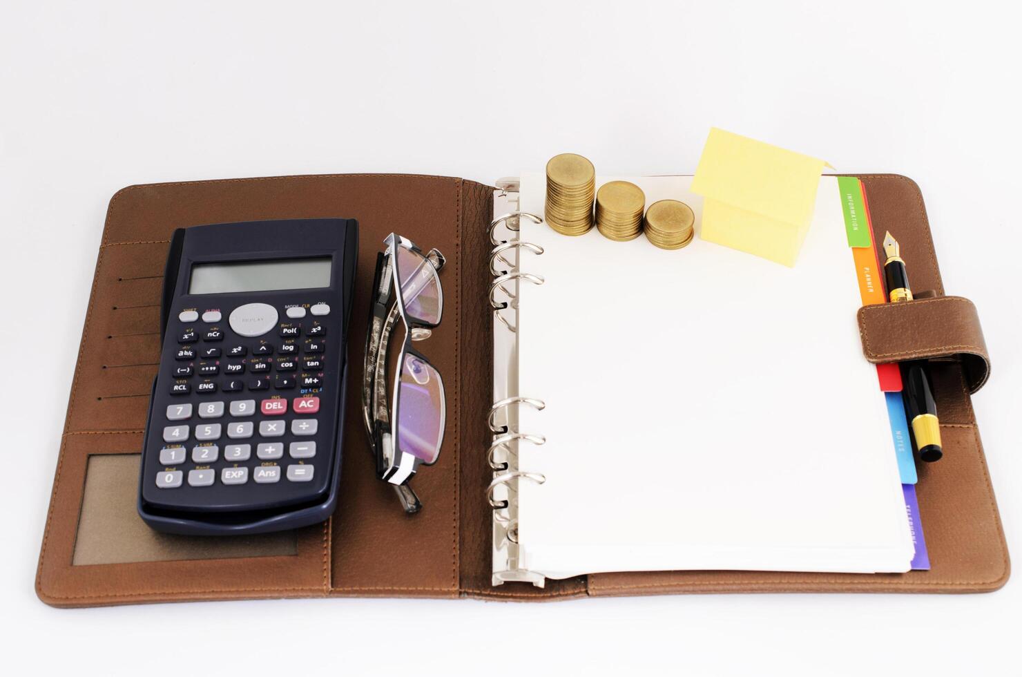 Geschäftskonzept mit Geldmünzenstapel und gelbem Papierhaus und Taschenrechner und Brille und Füllfederhalter auf Geschäftsmemobuch auf weißem Hintergrund foto