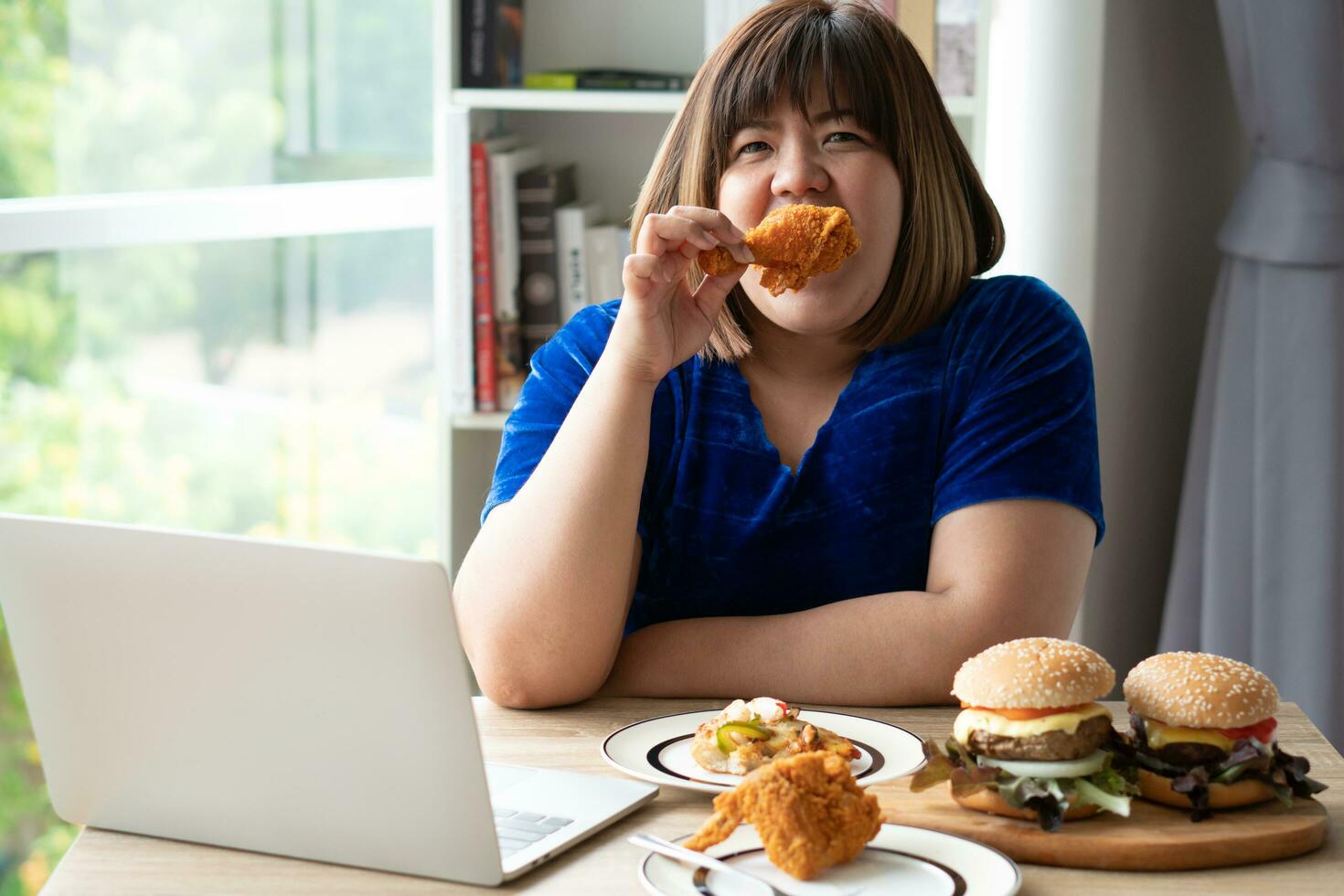 hungrig Übergewicht Frau halten gebraten Huhn, Hamburger auf ein hölzern Teller und Pizza auf Tisch, während Arbeit von heim, dazugewinnen Gewicht Problem. Konzept von Saufgelage Essen Störung Bett foto