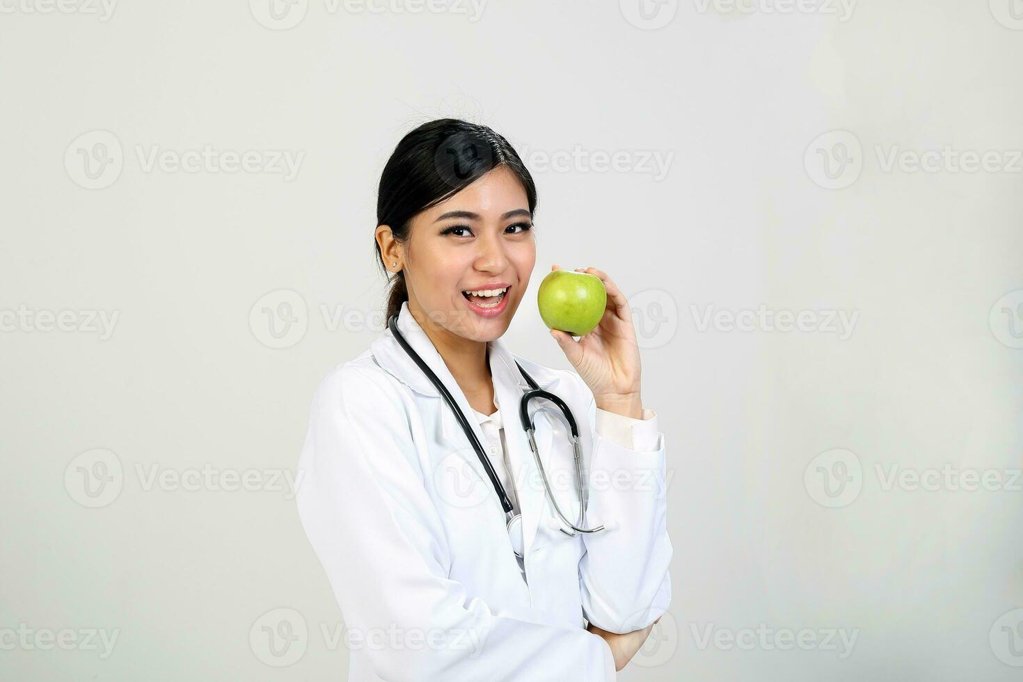 jung asiatisch weiblich Arzt tragen Schürze Uniform Tunika Stethoskop halten zeigen zeigen Essen gesund Grün Apfel foto