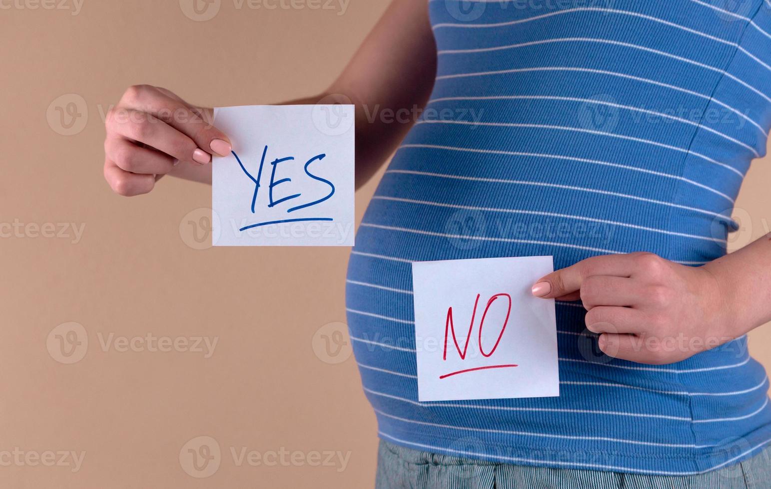 eine Nahaufnahme des Bauches einer schwangeren Frau in einem blauen T-Shirt, das zwei Blätter mit den Wörtern ja und nein hält foto