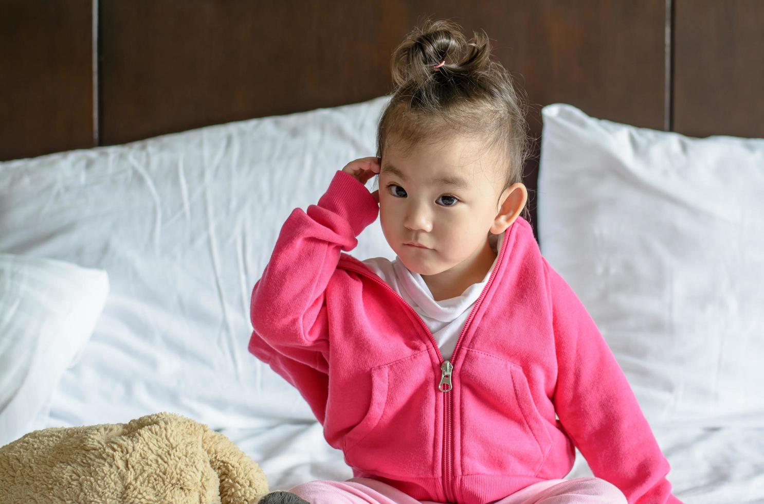 süßes asiatisches Baby, das eine rosa Jacke trägt, die auf dem Bett sitzt foto