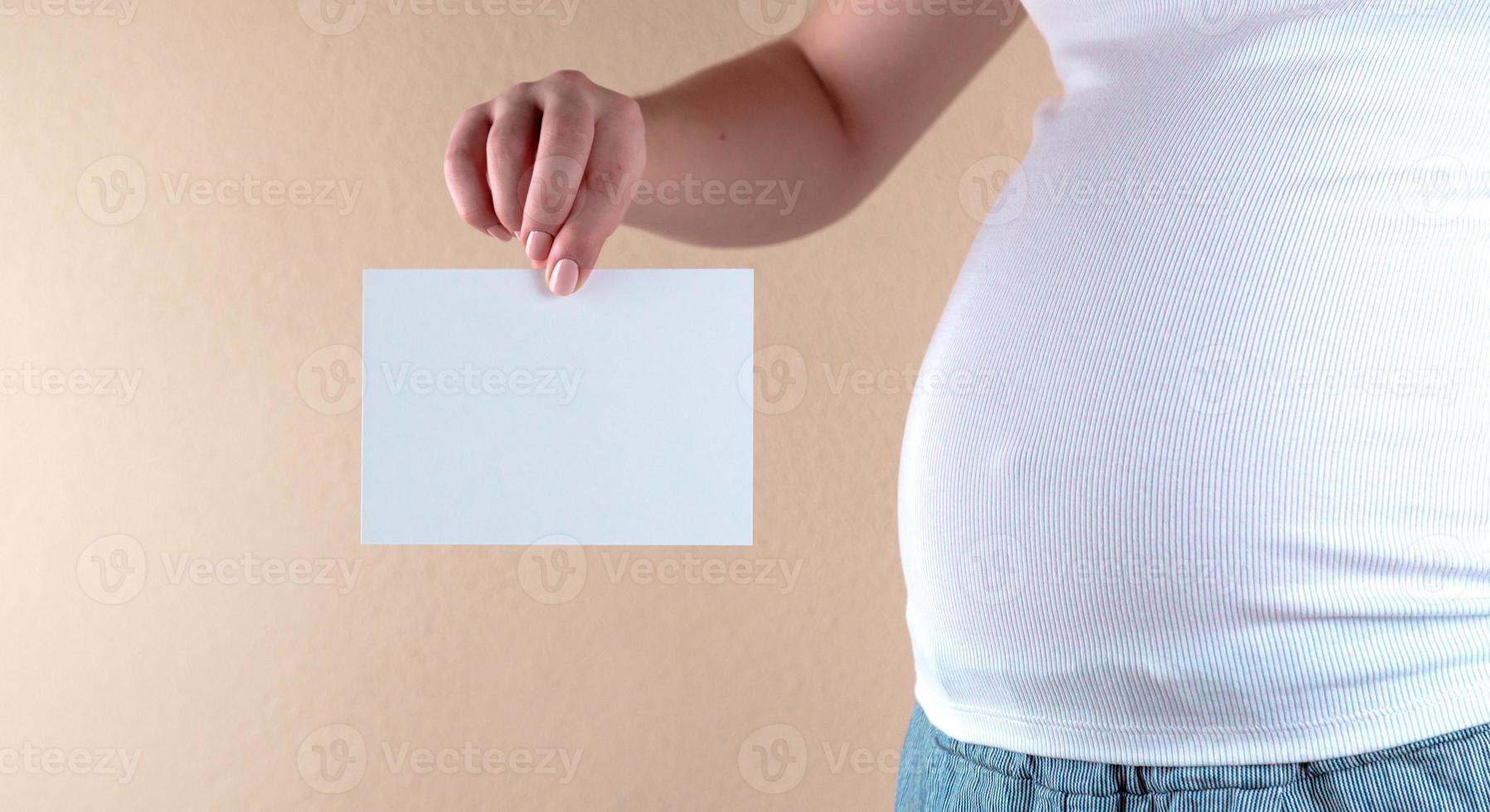 eine Nahaufnahme des Bauches einer schwangeren Frau, die ein leeres Blatt Papier hält foto