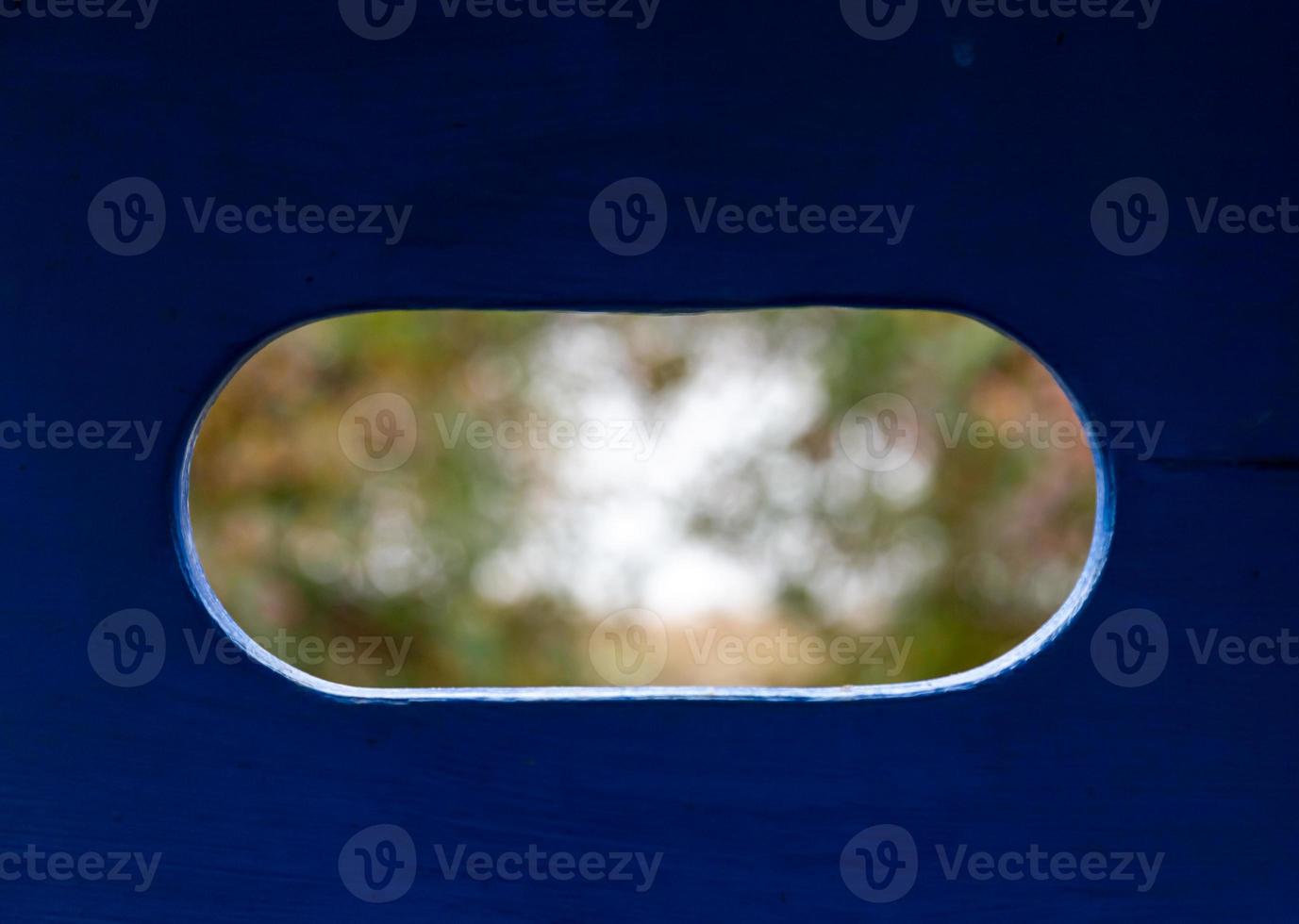 verschwommene Landschaftsansicht der Bäume im Park durch ein ovales Loch in einer blauen Holzwand foto