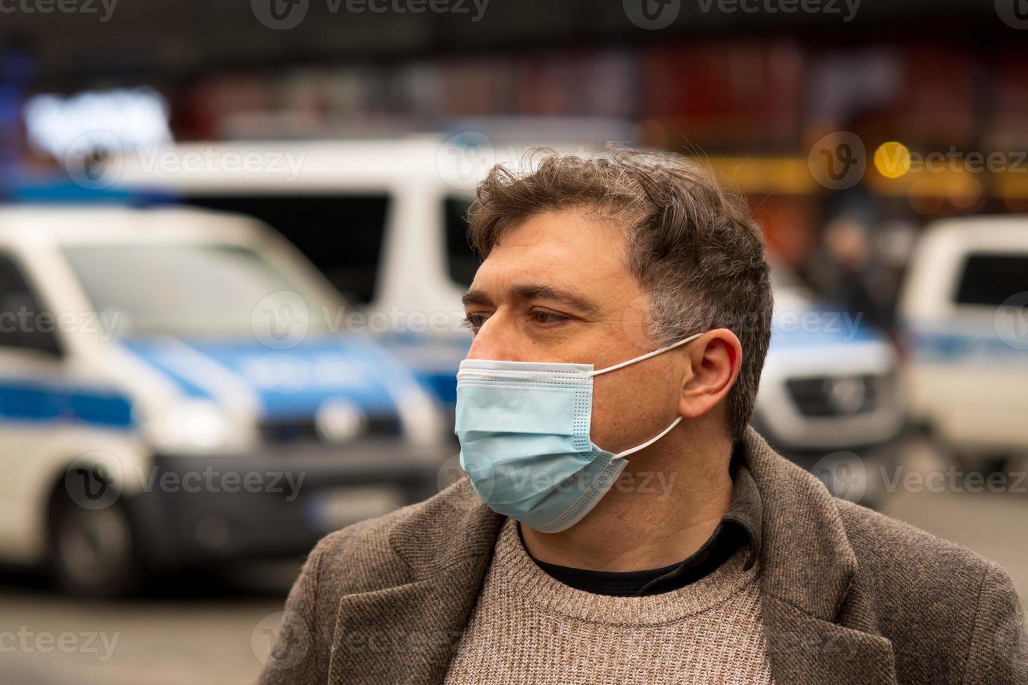 Außenporträt eines Mannes, der sein Gesicht mit einer medizinischen oder chirurgischen Maske schützt, die auf dem Hintergrund aus fokussierten Polizeiautos beiseite schaut foto