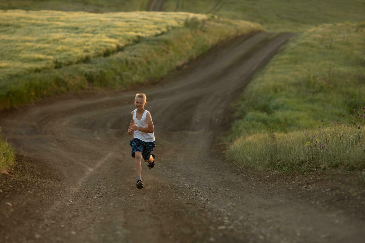 ein Junge im Weiß T-Shirt läuft beim Sonnenuntergang entlang ein ländlich Straße Nächster zu ein Weizen Feld. Sommer- Ferien im das Landschaft. glücklich Kindheit foto