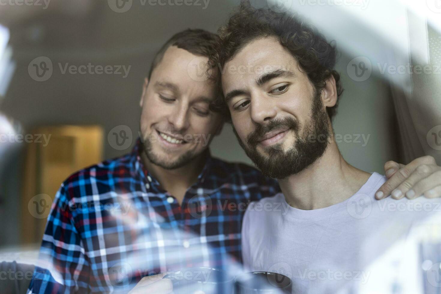 jung Fröhlich Paar im Liebe suchen aus das Fenster. zwei jung androgyn Männer lächelnd zusammen und haben Kaffee. foto