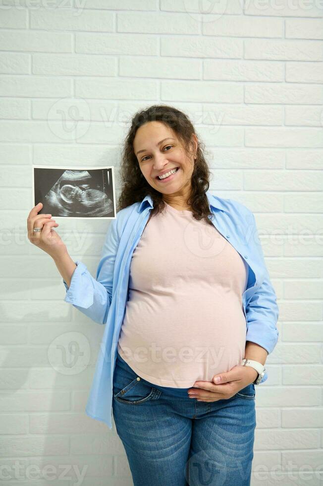 glücklich positiv Erwachsene schwanger Frau, streicheln ihr Bauch, lächelnd, zeigen beim Kamera das Ultraschall von ihr Baby im Gebärmutter foto