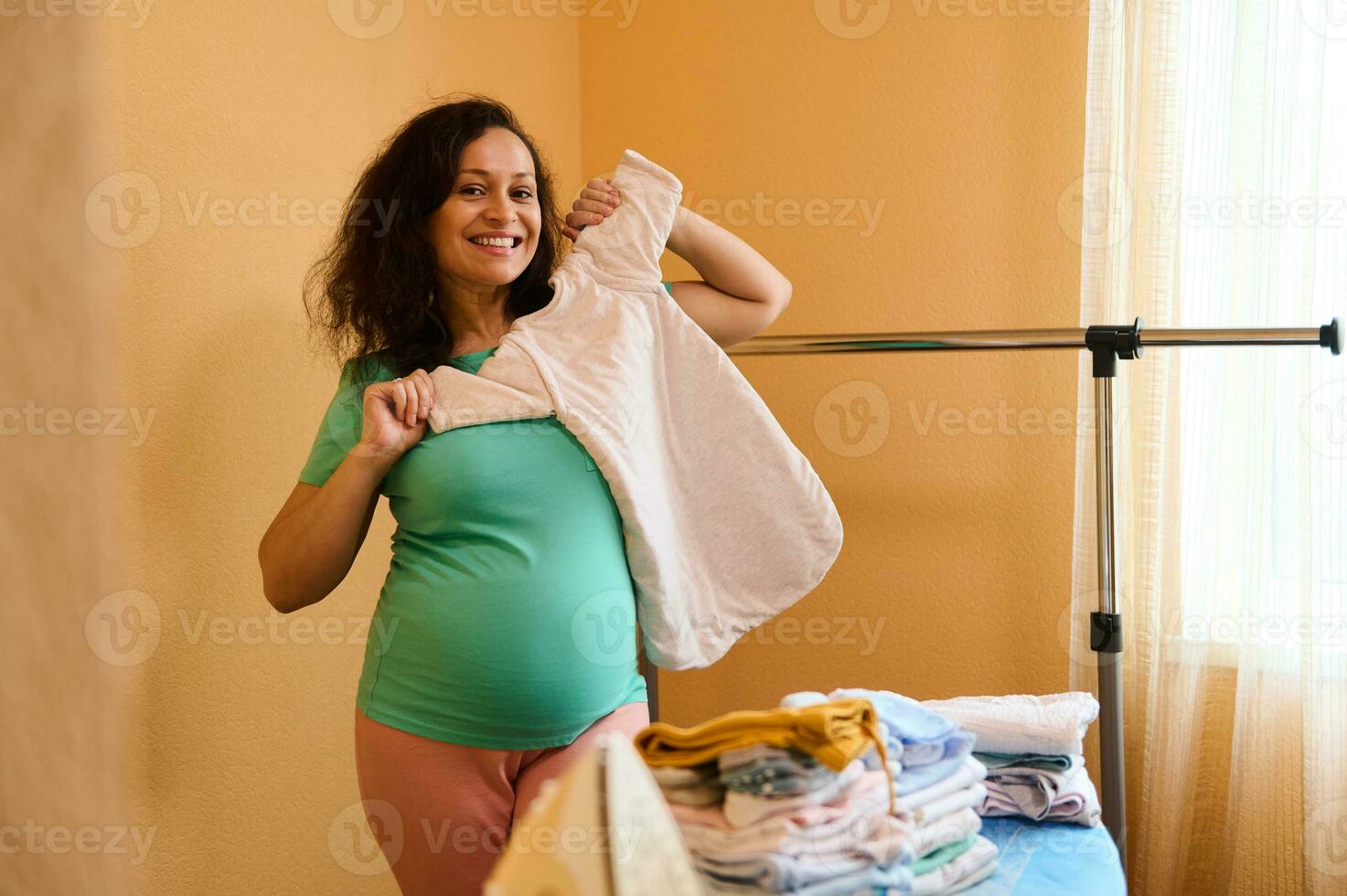 schön glücklich schwanger Frau halten ein Schlafen Tasche zum ihr Zukunft Neugeborene Baby, Stehen beim das Bügeln Tafel beim Zuhause foto