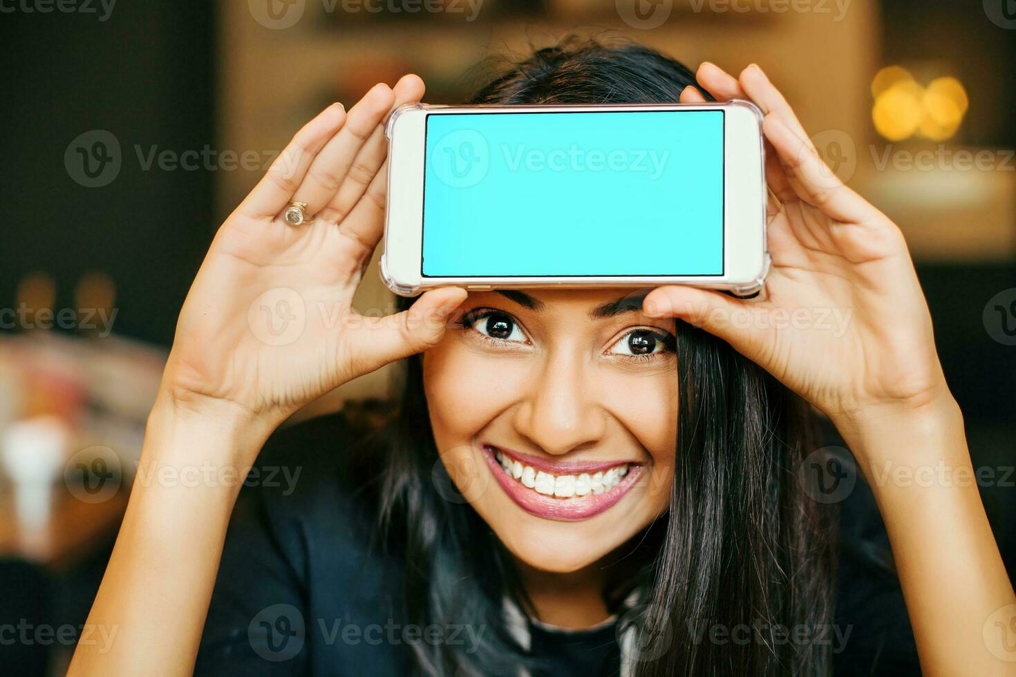 ein lächelnd jung indisch Frau posieren mit ihr Telefon gehaltenen zu ihr Stirn foto